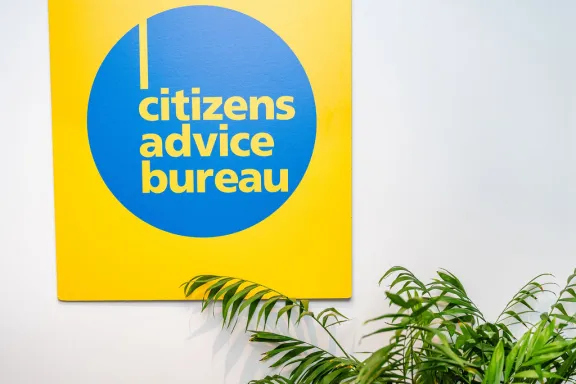 A yellow and Blue sign on a white wall saying Citizens Advice Bureau with some plants at the bottom